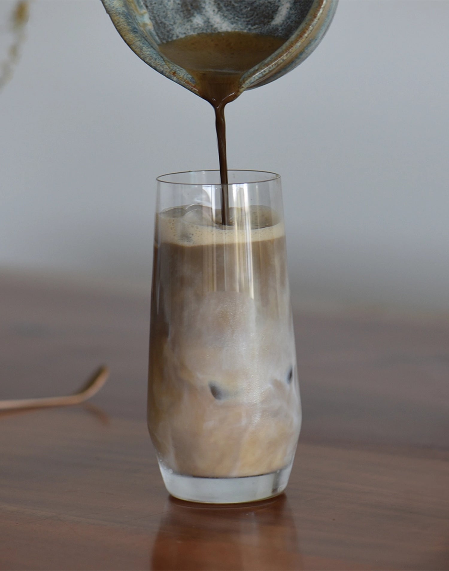 Hot liquid being poured into a glass of iced coffee on a wooden surface.