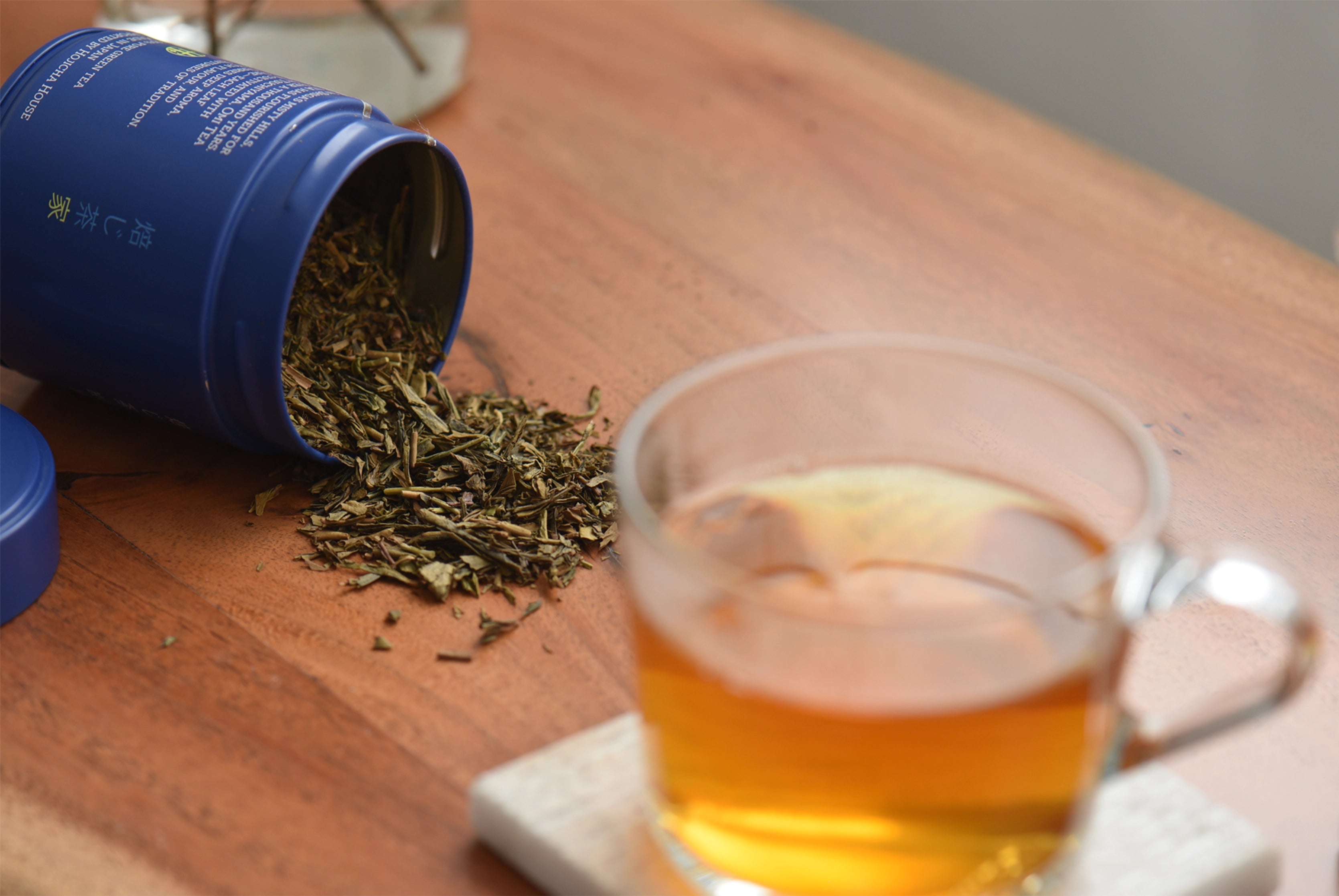 Tea leaves spilling from a blue container onto a wooden surface with a glass of tea in the foreground.